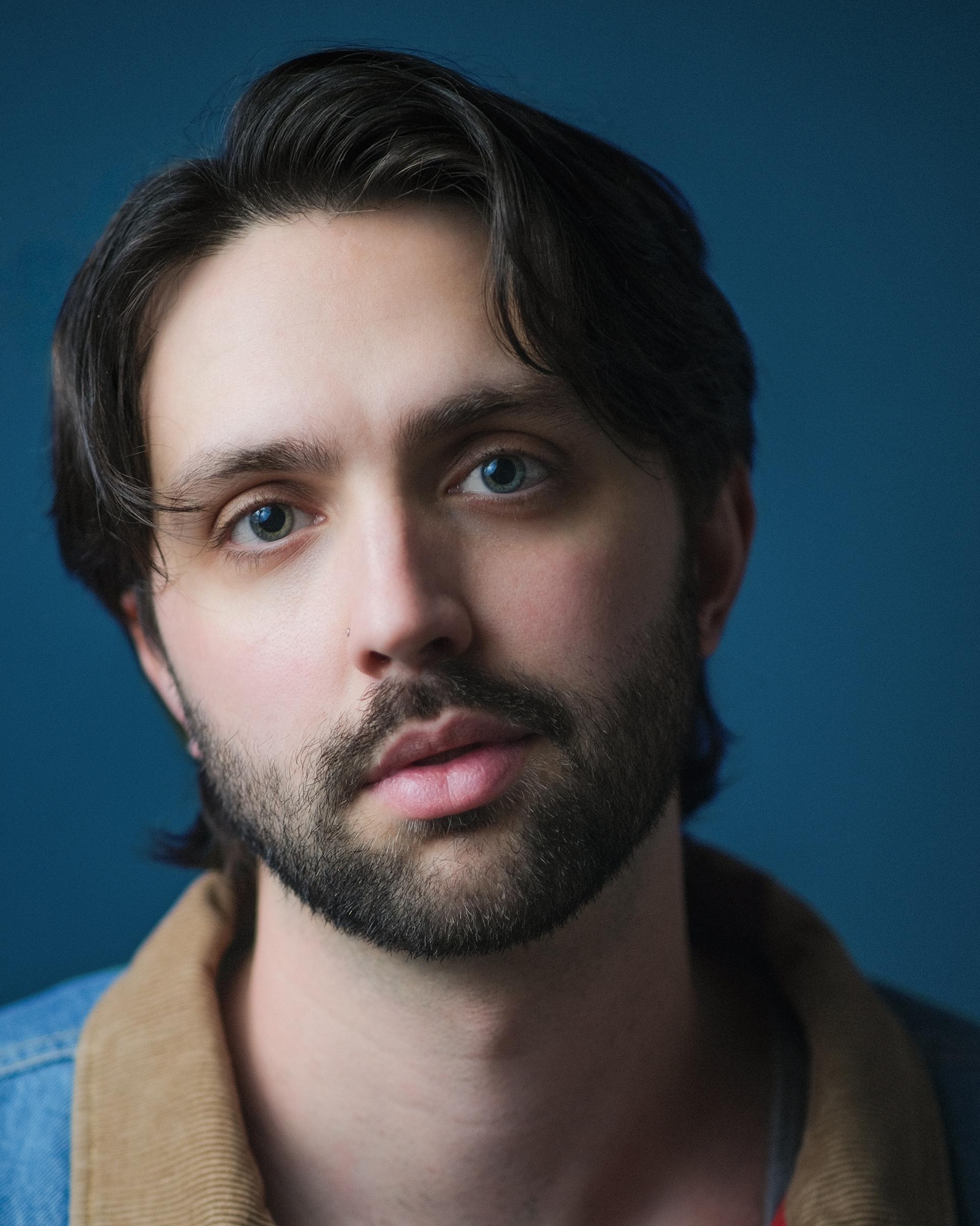 Headshot of Shane Lonergan in a tan jacket against a blue-grey background